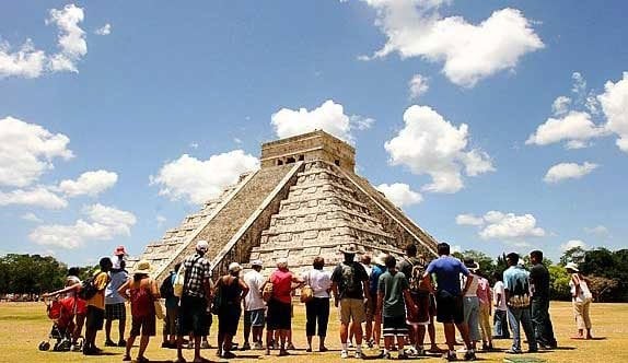 Turistas frente a la pirámide maya de Chichén Itzá bajo un cielo azul con nubes