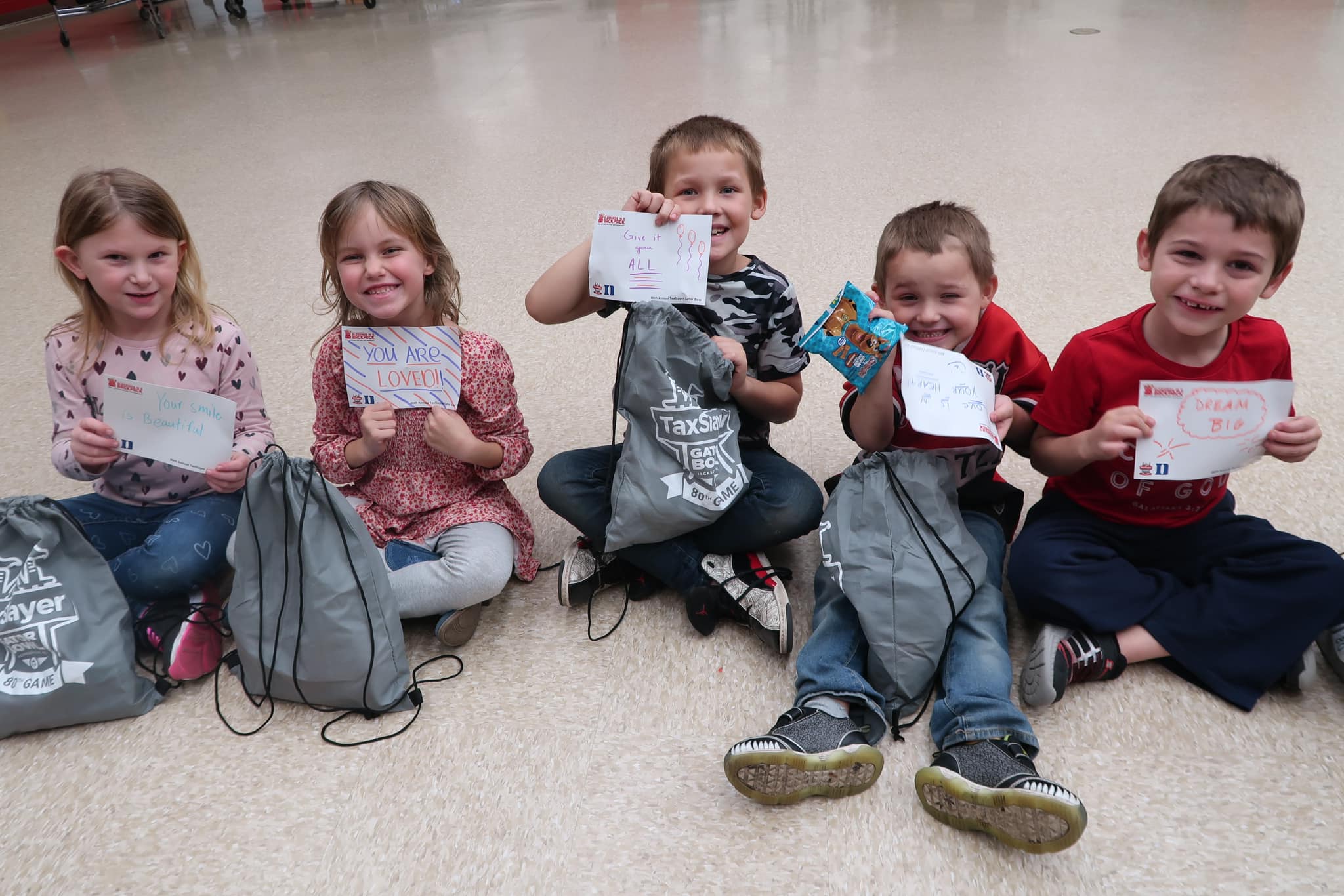 Smiling child with a newspaper hat in a classroom setting, surrounded by other children in school uniforms engaged in activities.