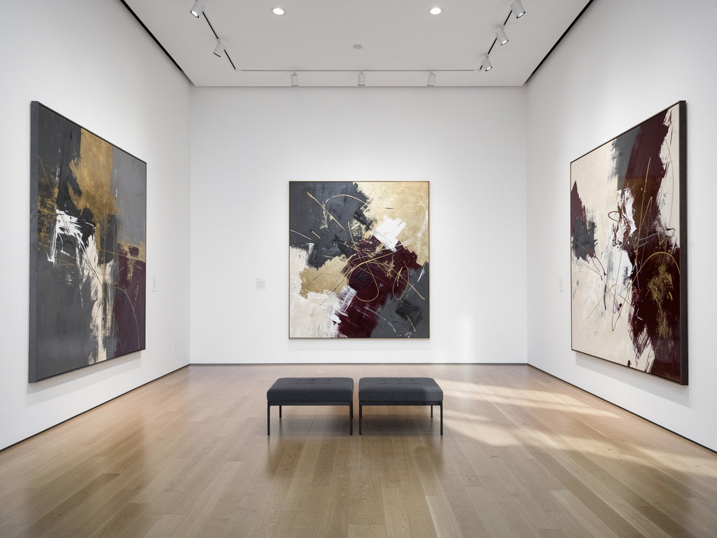 Galería de arte moderno con cuadros abstractos grandes, bancos negros y suelo de madera