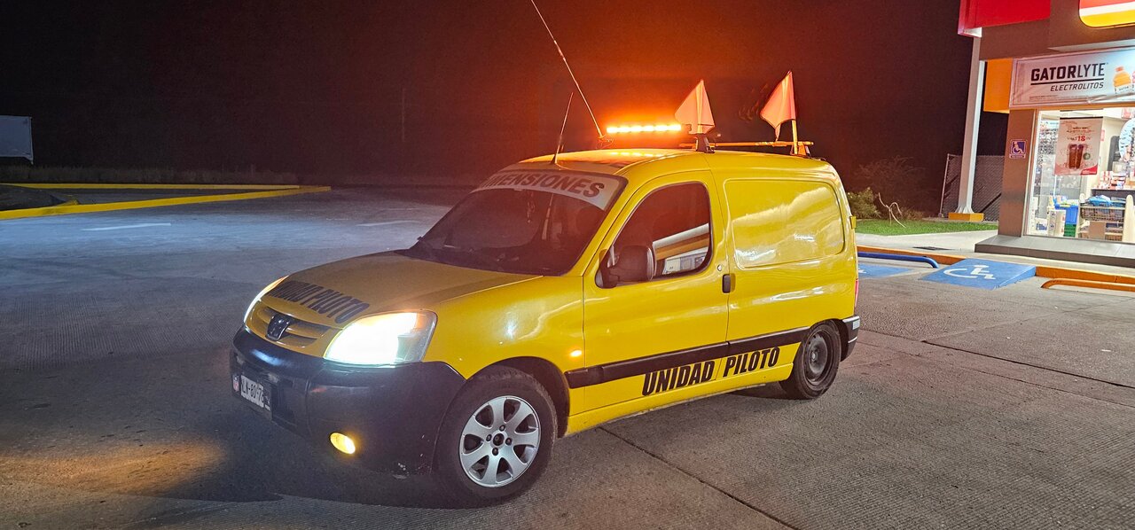 Furgoneta amarilla de unidad piloto con luces encendidas estacionada en una gasolinera durante la noche.