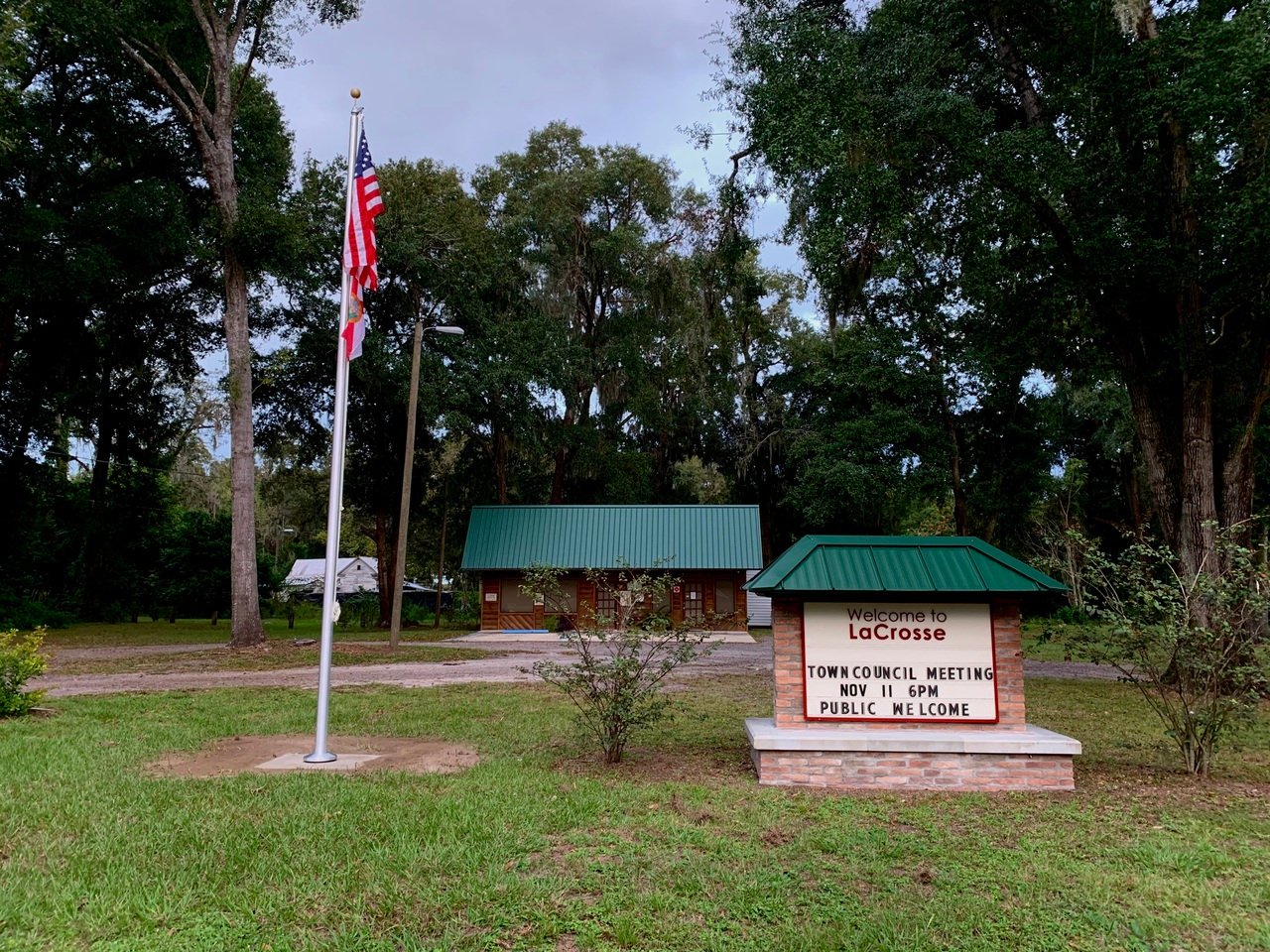 Entrance sign to LaCrosse town with meeting details, surrounded by green trees and an American flag in the foreground.