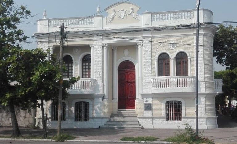 Fachada de una casa antigua de estilo colonial con columnas y puerta roja en una calle tranquila, rodeada de árboles.