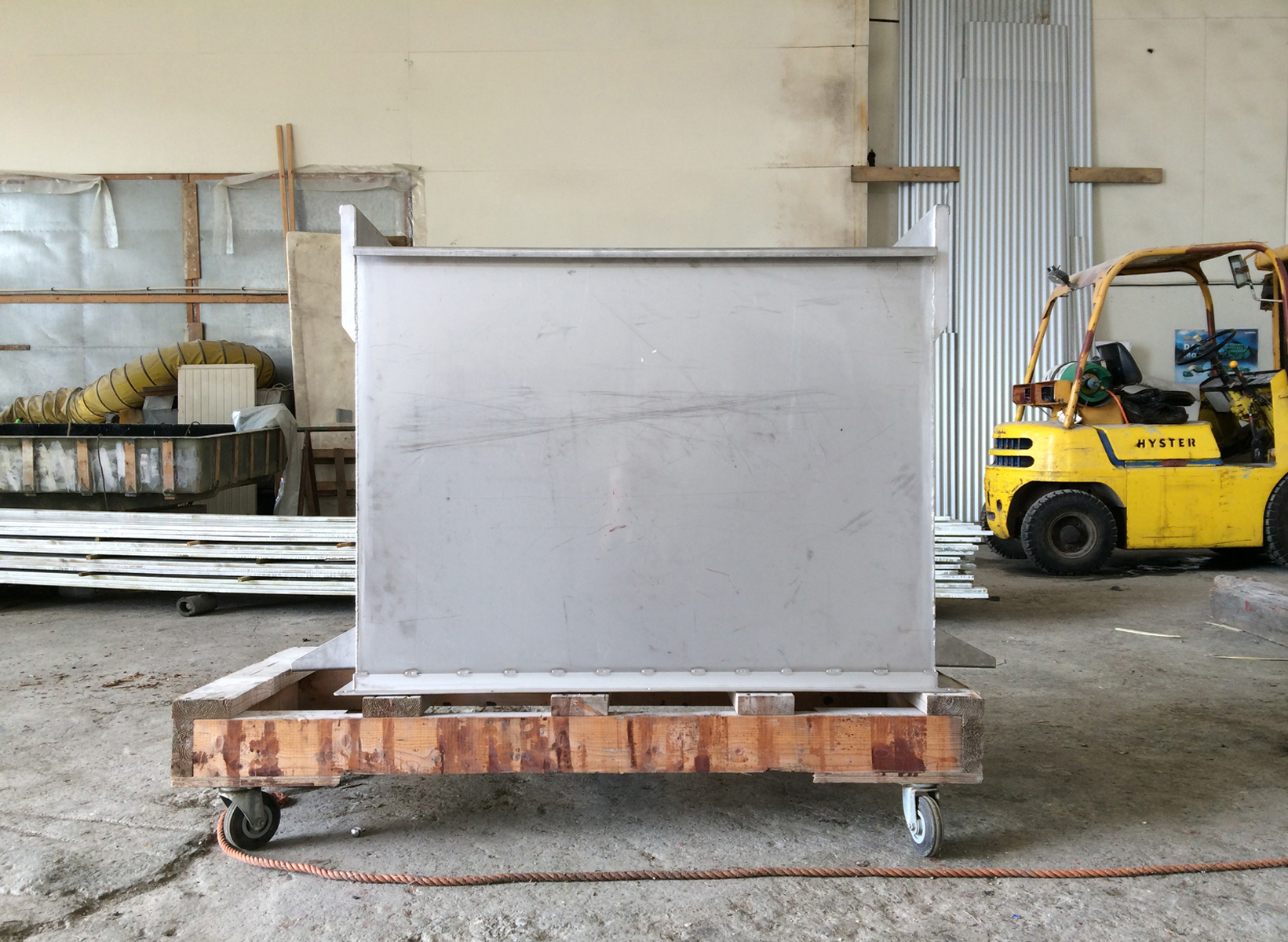 A bare aluminum box sits on a pallet dolly in a warehouse with a yellow forklift.