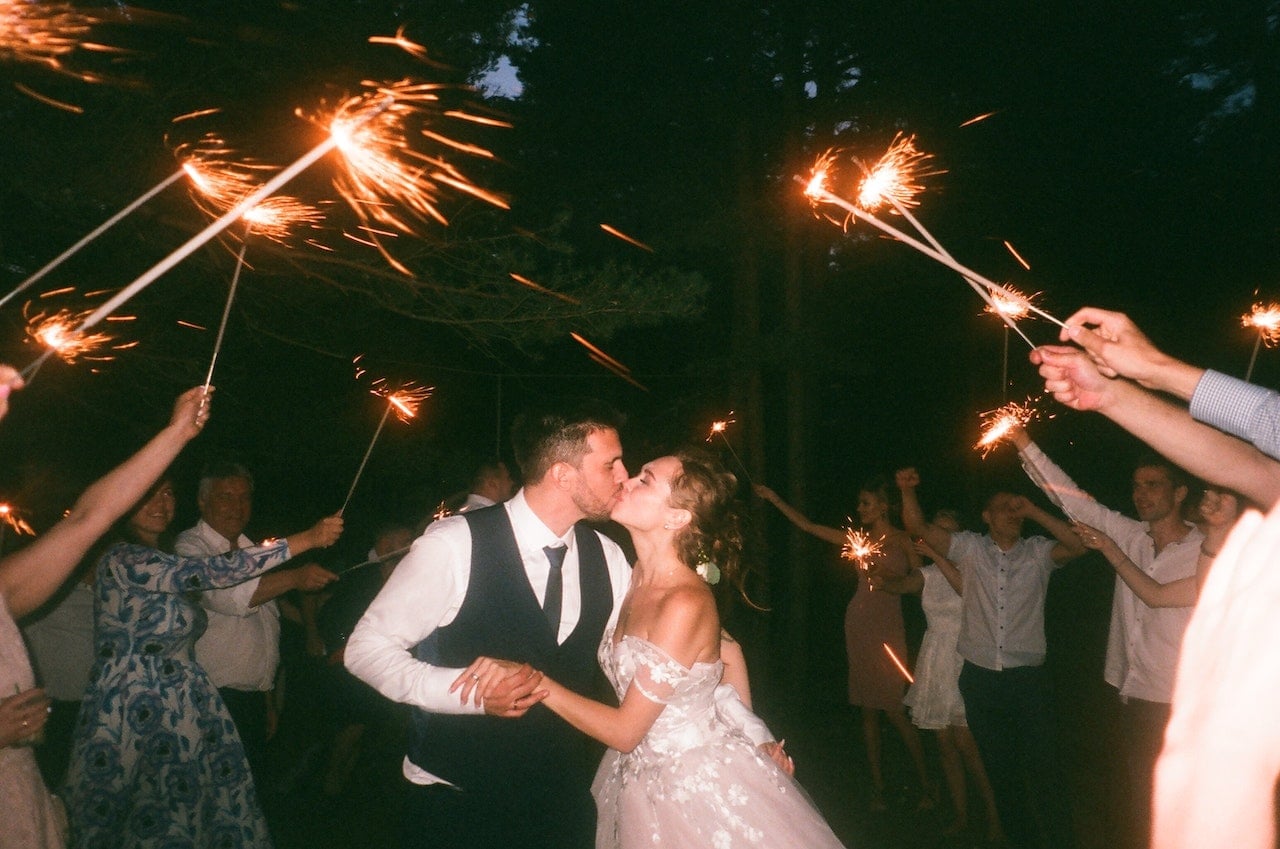 Bride and groom kiss surrounded by guests holding bright sparklers at night wedding celebration.