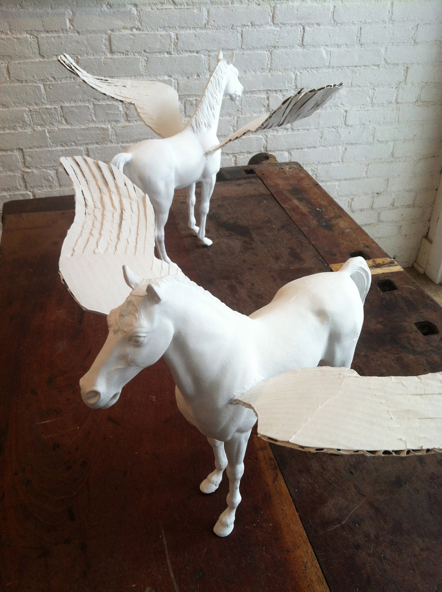 A pair of model horses with cardboard wings, all painted white, on a workbench.