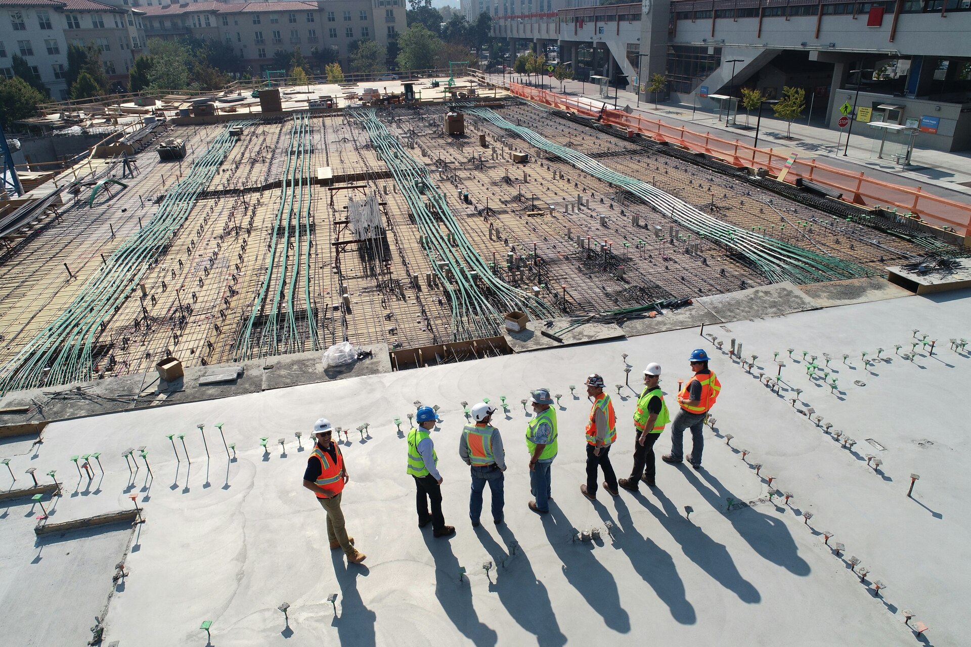 A group of construction workers in safety gear standing on a large building site with foundations and rebar visible.