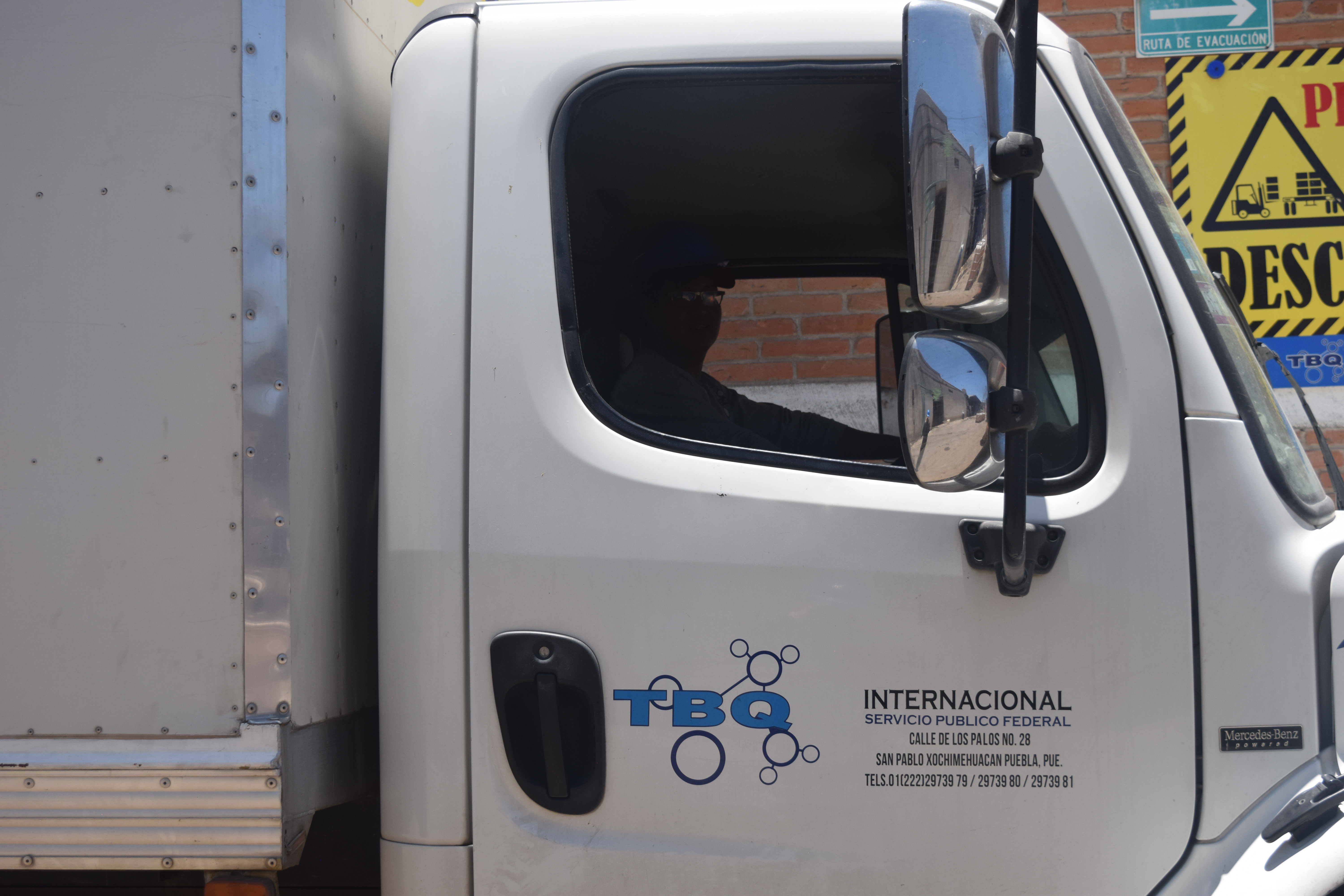 Trabajadores con cascos y chalecos de seguridad frente a camionetas en una empresa de transporte con cartel de TBQ Internacional.