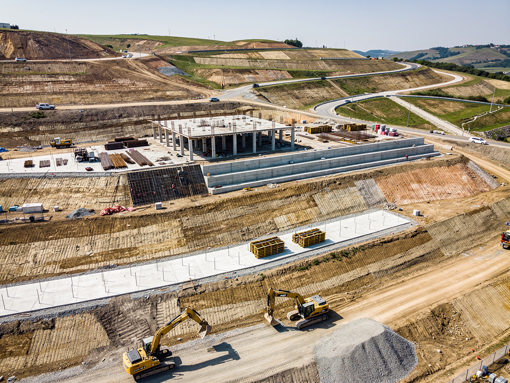 Vista aérea de obra de construcción de carretera con excavadoras, estructuras de hormigón y taludes en terreno montañoso