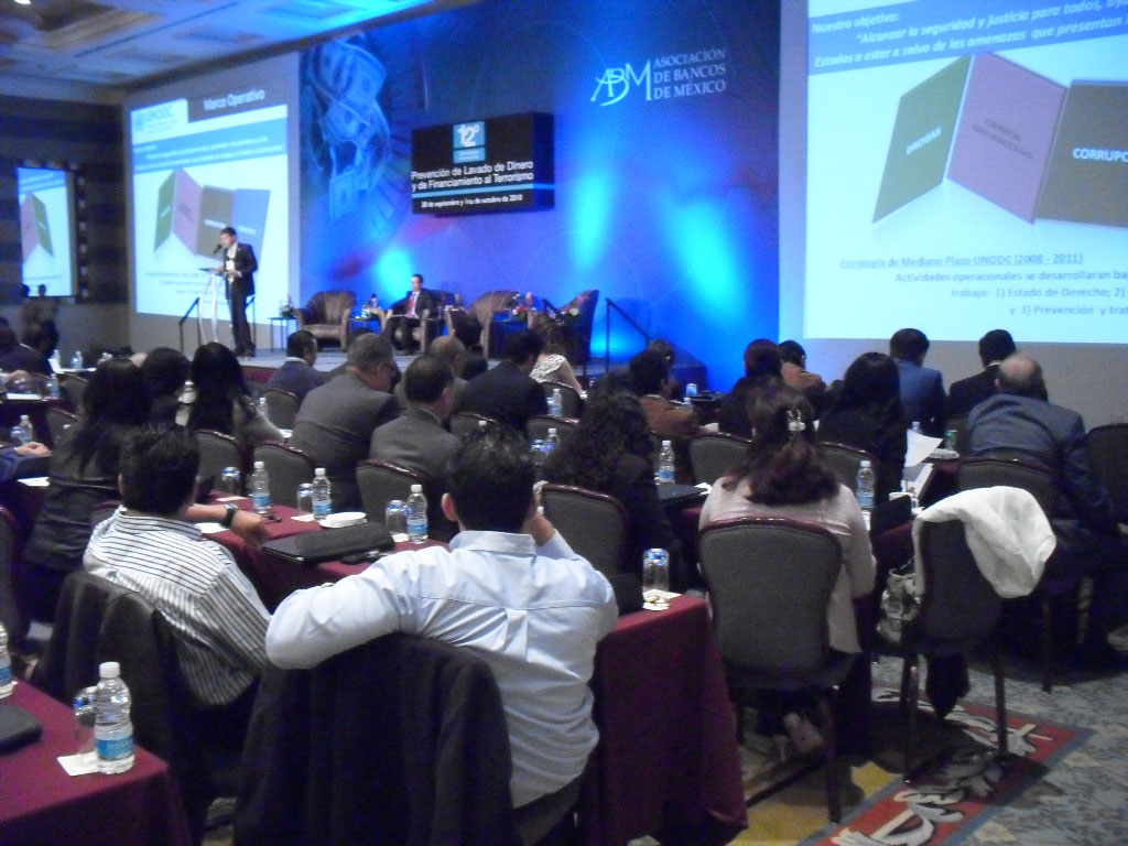 Conferencia en curso sobre la prevención de lavado de dinero, con asistentes sentados y un presentador frente a una pantalla grande en un salón de la Asociación de Bancos de México.