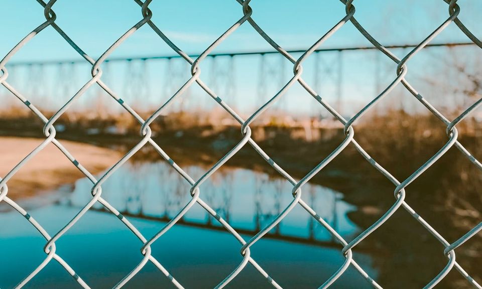 Vista de un río y puente de ferrocarril a través de una cerca de malla metálica bajo un cielo azul claro.