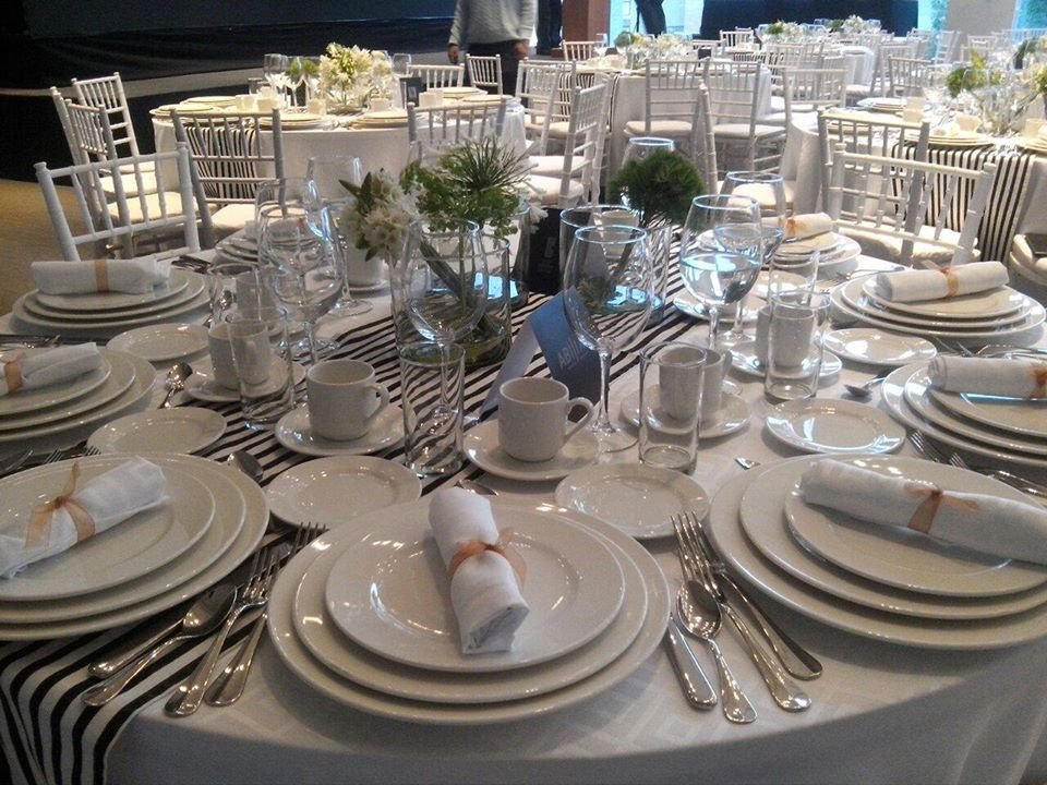 Mesa redonda elegantemente decorada con platos blancos, cubiertos de plata, copas de cristal y servilletas blancas, rodeada de sillas blancas y centros de mesa florales.