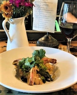 Elegant dish with shrimp and greens served on a white plate, accompanied by a wine glass and a flower vase on a wooden table.
