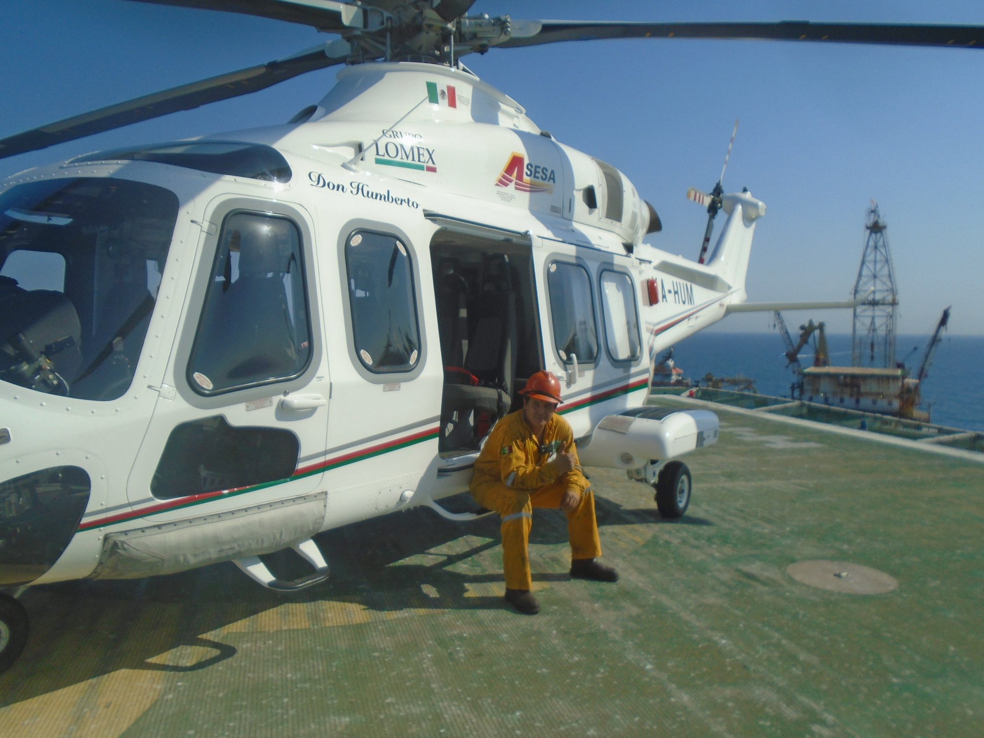 Trabajador con casco descansando al lado de un helicóptero en una plataforma marítima con vista al océano y estructuras petroleras.
