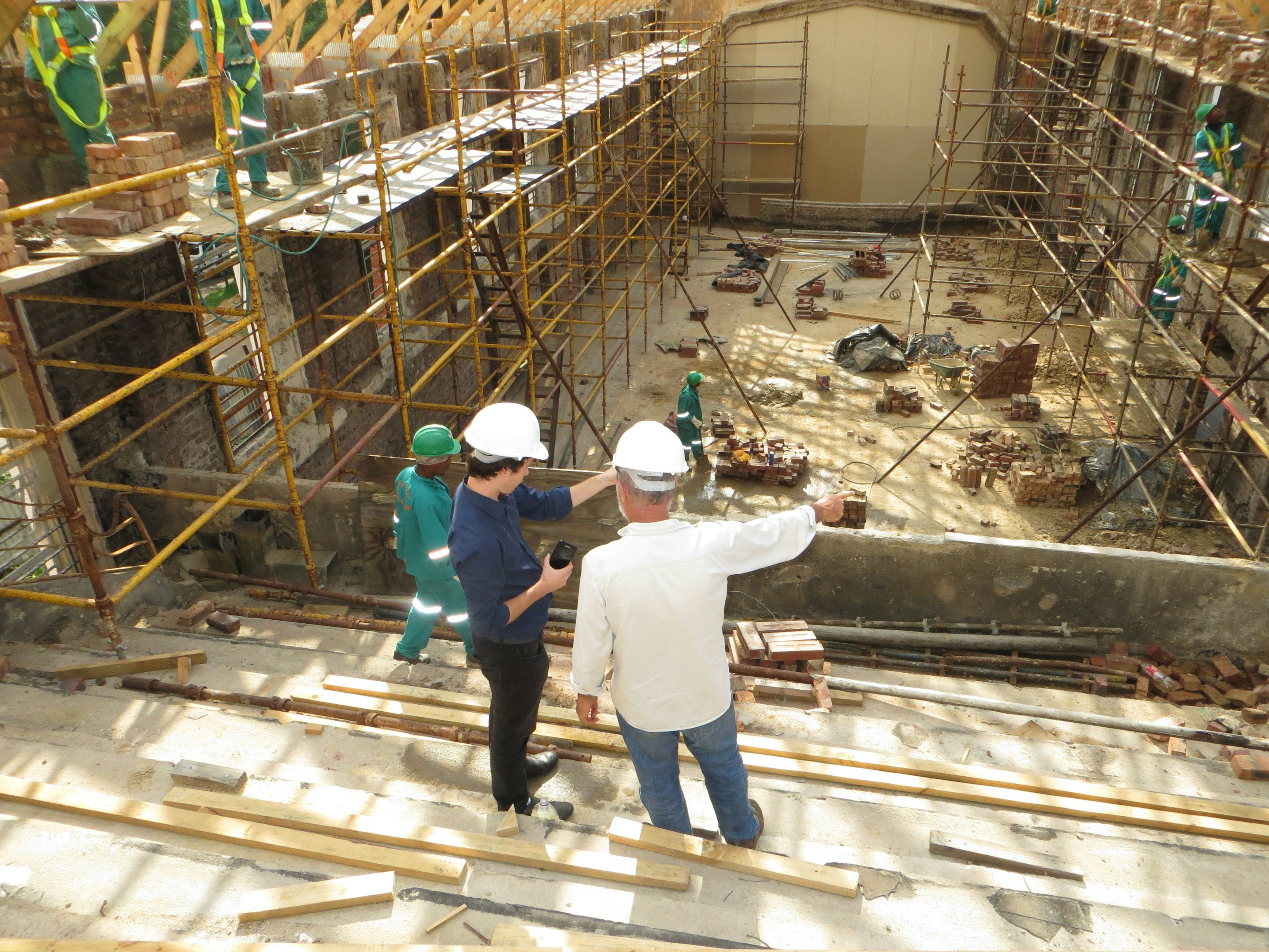 Construction site with workers on scaffolding, two supervisors wearing hard hats inspecting progress, and building materials scattered on the ground.