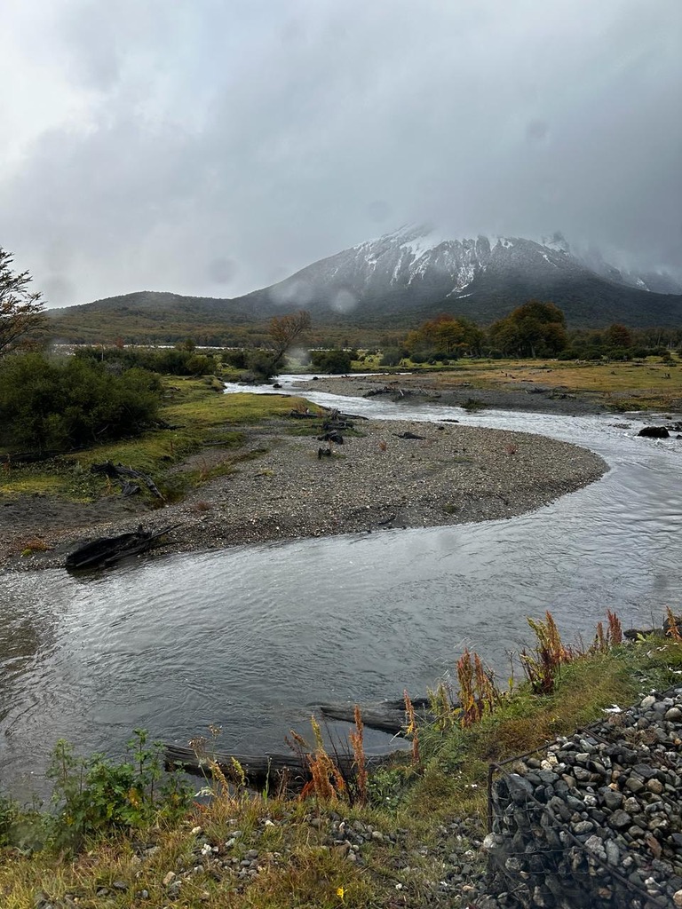 Paisaje montañoso con río serpenteante y montaña nevada al fondo bajo un cielo nublado.