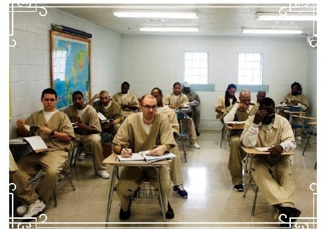 Inmates attending a classroom session in a prison setting, wearing beige uniforms, seated at individual desks with notebooks and educational materials.