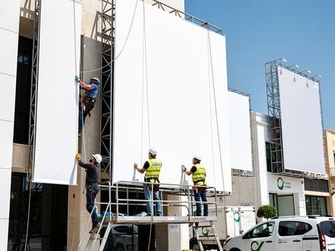Trabajadores instalando grandes paneles blancos en la fachada de un edificio comercial con plataforma elevadora