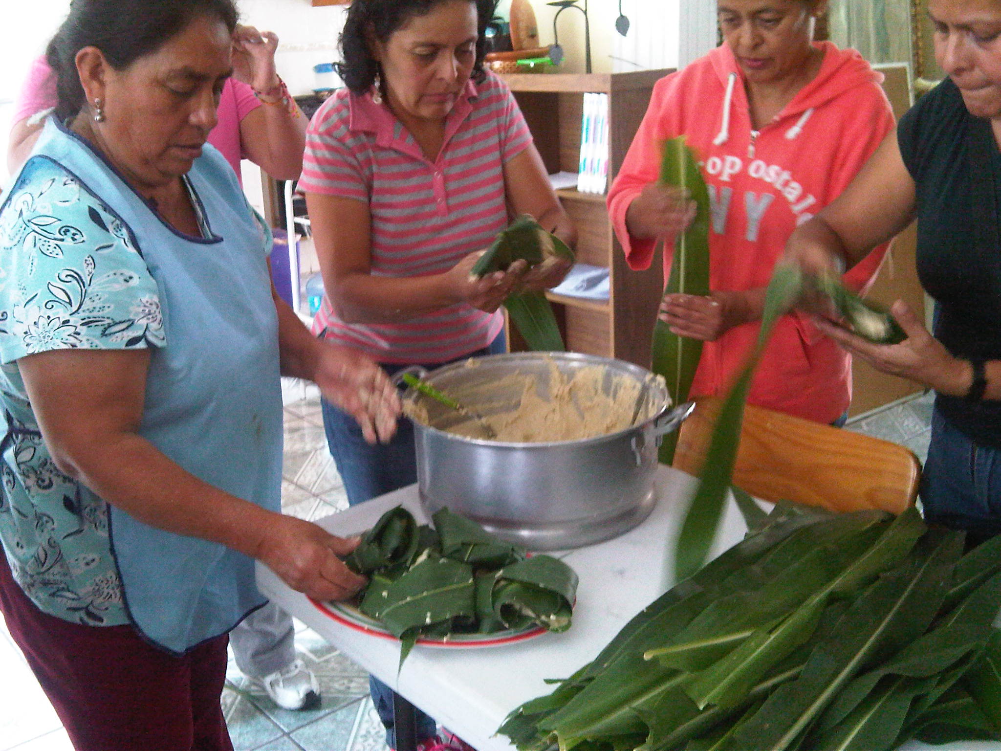 Un grupo de mujeres preparando tamales en hojas verdes sobre una mesa con masa en una olla.