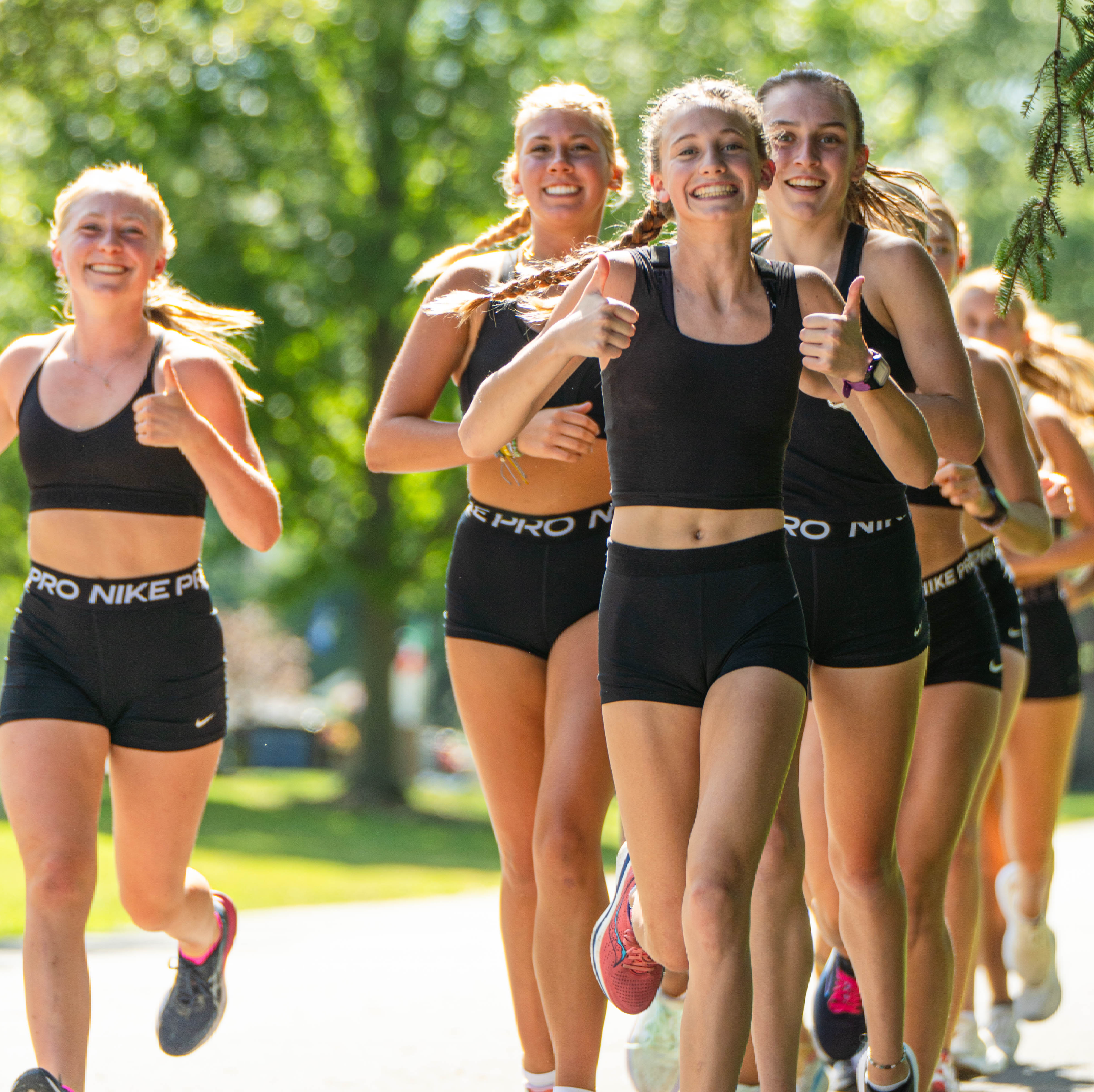 Group of young women jogging in a park, wearing athletic wear, giving thumbs up and smiling, with trees in the background.