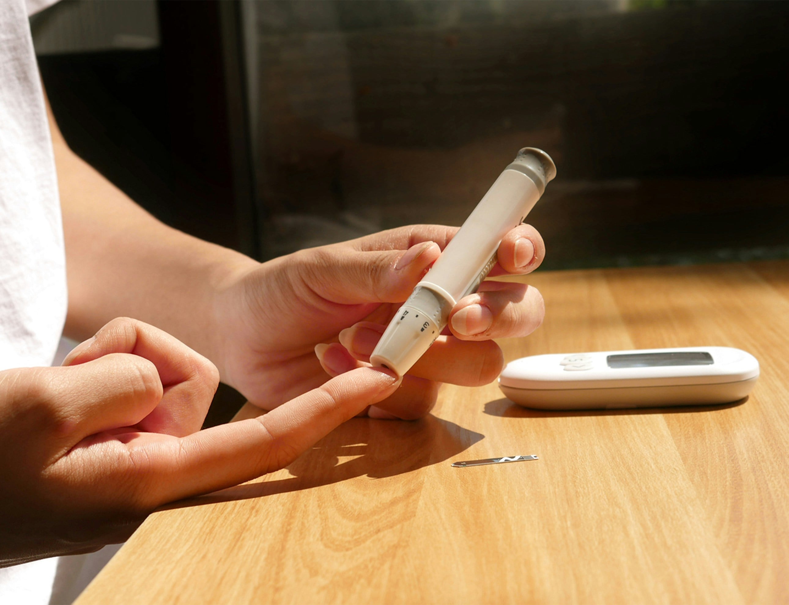 Person using a blood glucose meter lancet pen to check blood sugar at a wooden table