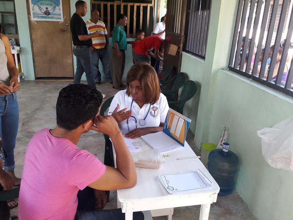 Persona recibiendo consulta médica en centro de salud comunitario, con doctora realizando evaluación y más personas en fila al fondo.