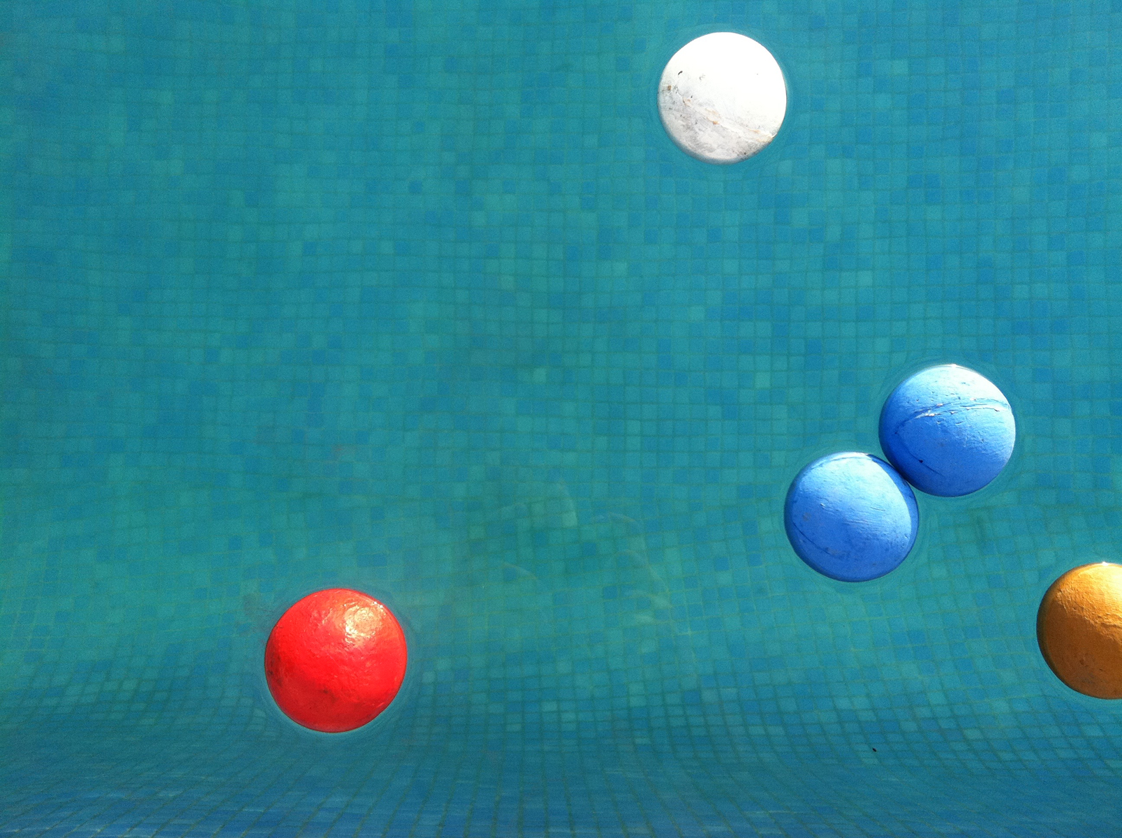 Five painted balls, blue, red, yellow and white, float in a sunny, blue-tiled pool.