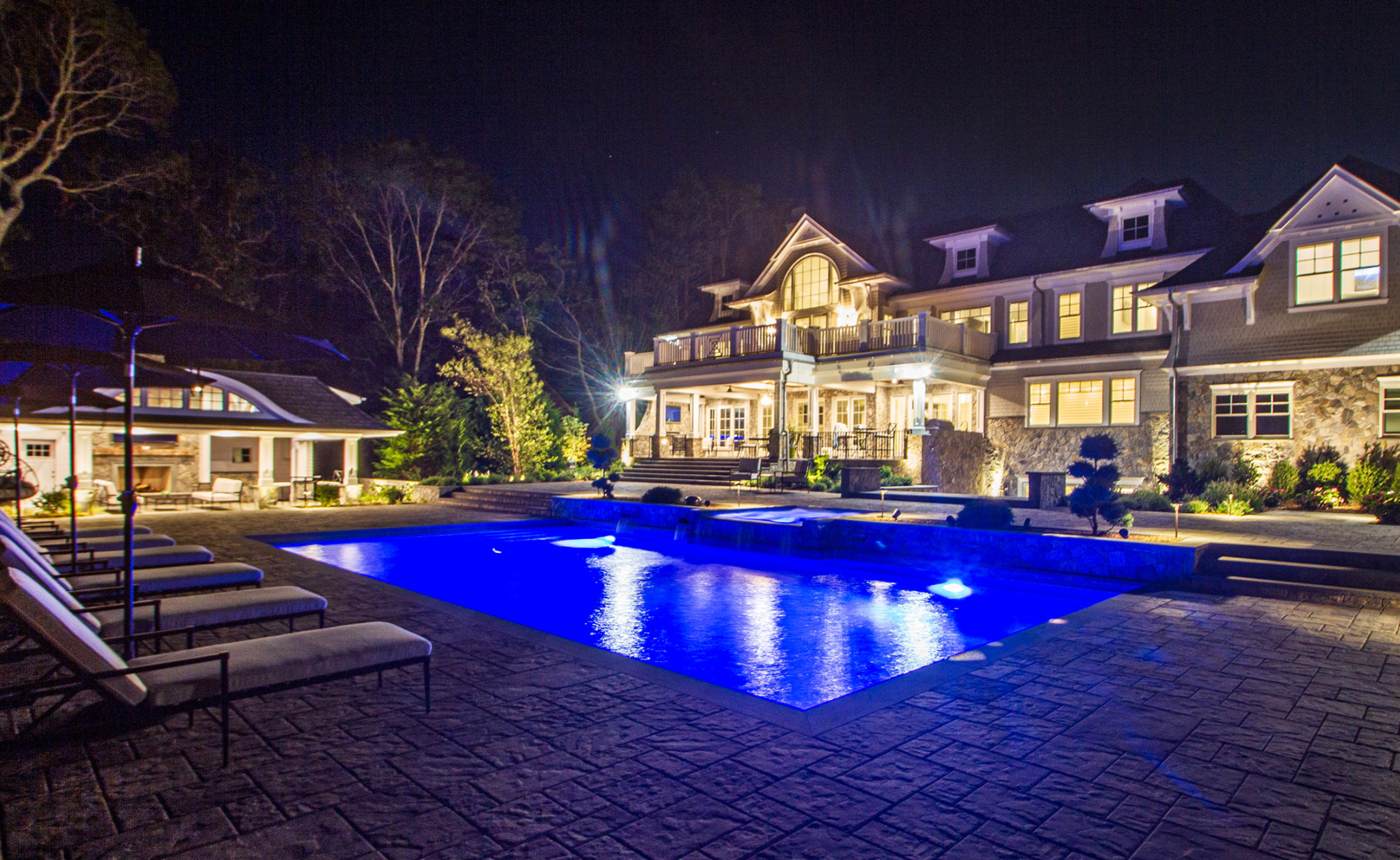 Luxurious mansion exterior at night with illuminated swimming pool, landscaped garden, and multiple lounge chairs.