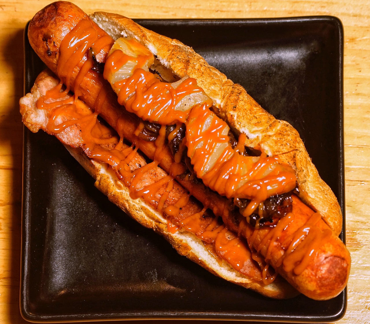 Gourmet hot dog in a bun topped with grilled onions and drizzle of orange sauce on a black plate.
