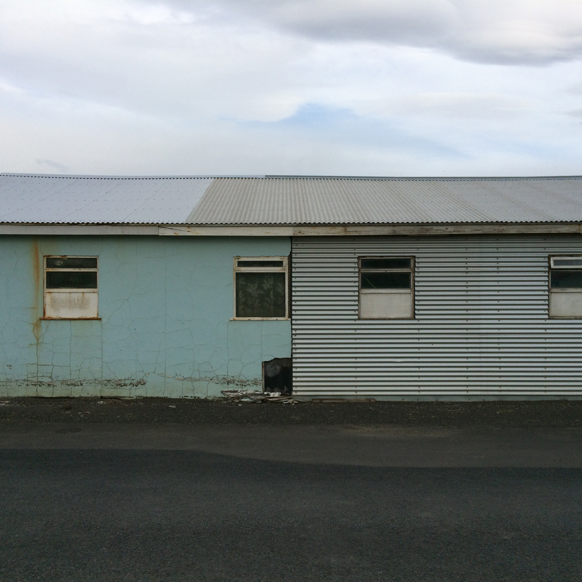 A light blue and grey corrugated metal-roofed building with four small windows.