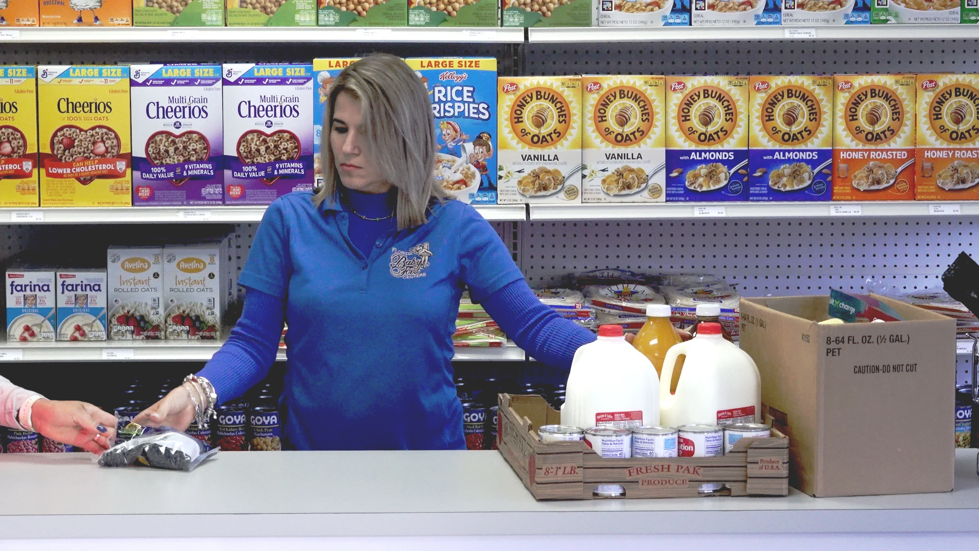 Two grocery store employees in green aprons managing produce and products at a counter, with various fruits and packaged items visible, in a well-lit interior.
