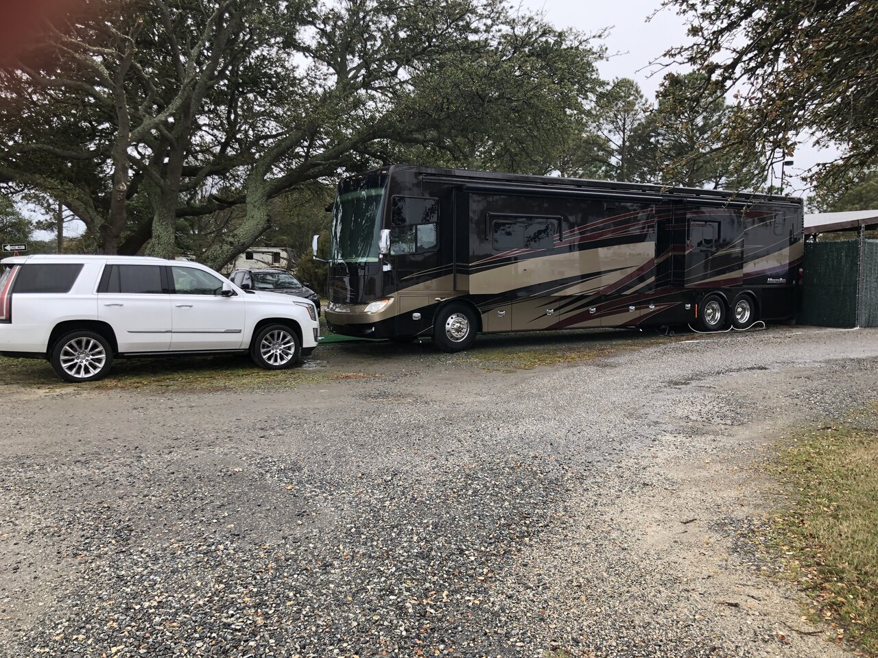 A large black and beige motorhome parked beside a white SUV on a gravel lot with trees in the background.