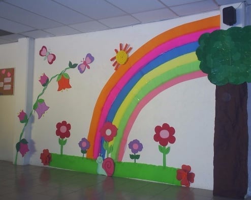 Decoración de pared con arcoíris de colores, flores, árbol y mariposas en aula escolar.