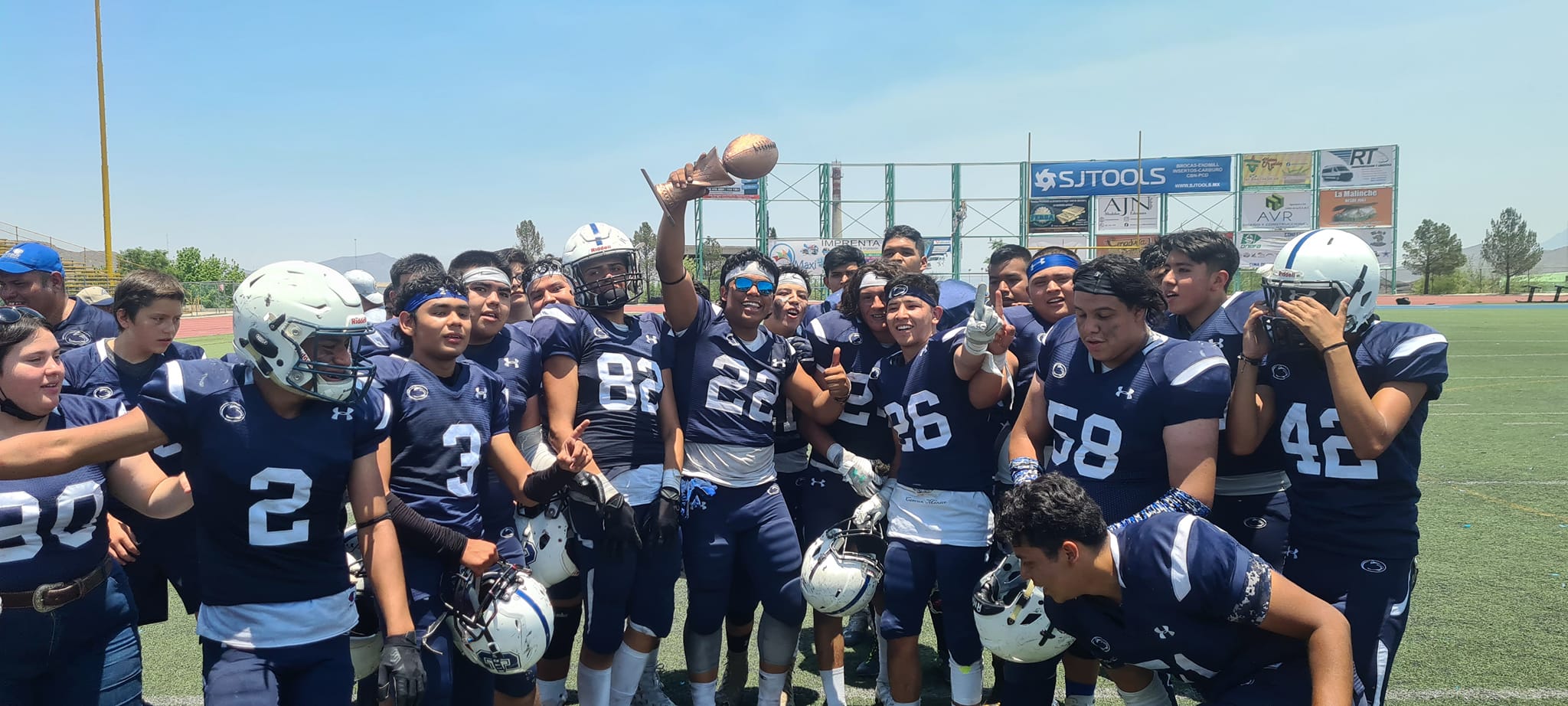 Equipo de fútbol americano juvenil celebrando con un balón en un campo deportivo bajo un cielo despejado.