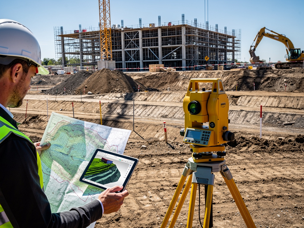Topógrafo con casco revisa planos y modelo 3D junto a estación total en obra de construcción con grúa y excavadora