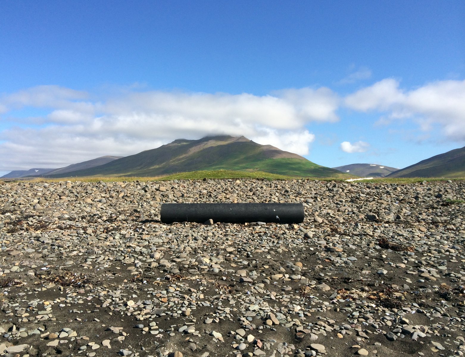 A black plastic tube on a gravel beach with mountains behind, and a sunny sky.