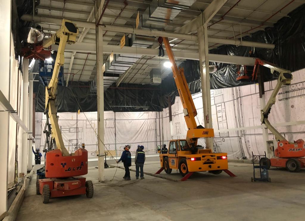 Excavadora amarilla en un almacén industrial con suelo brillante, plantas y ventanas grandes.