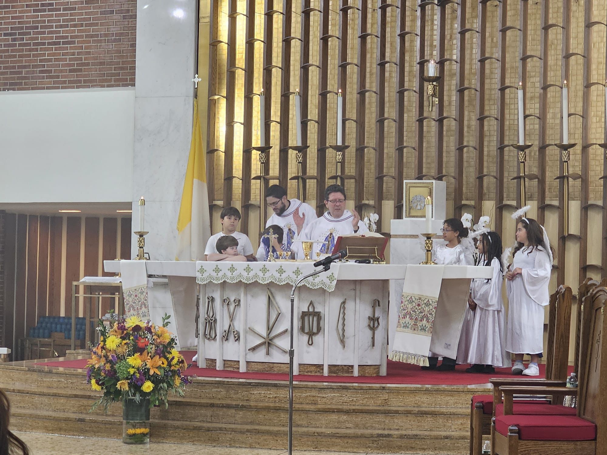 Priests conducting a church service at an altar with children in white robes, surrounded by ornate wooden decor and floral arrangements.