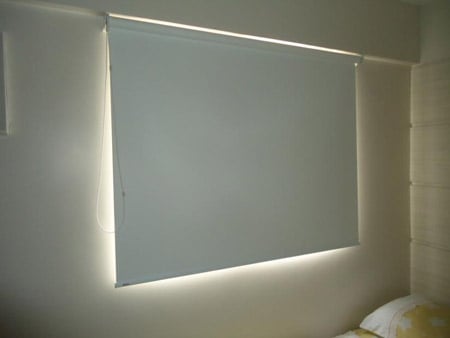 Persiana rolo cinza fechada em janela de quarto moderno, com cama parcialmente visível.