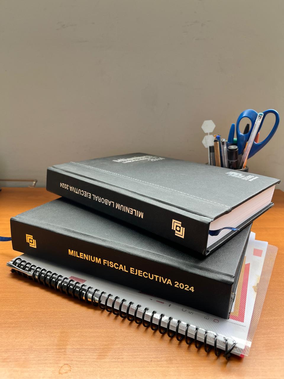 Libros 'Mileniun Fiscal Ejecutiva 2024' y 'Mileniun Laboral Ejecutiva 2024' apilados sobre un escritorio con una libreta, un portapapeles y portabolígrafos con tijeras y bolígrafos.