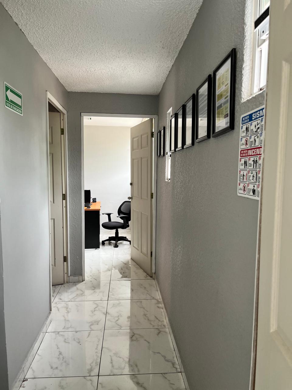 Pasillo interior con señal de evacuación y puerta abierta mostrando oficina con silla y escritorio.