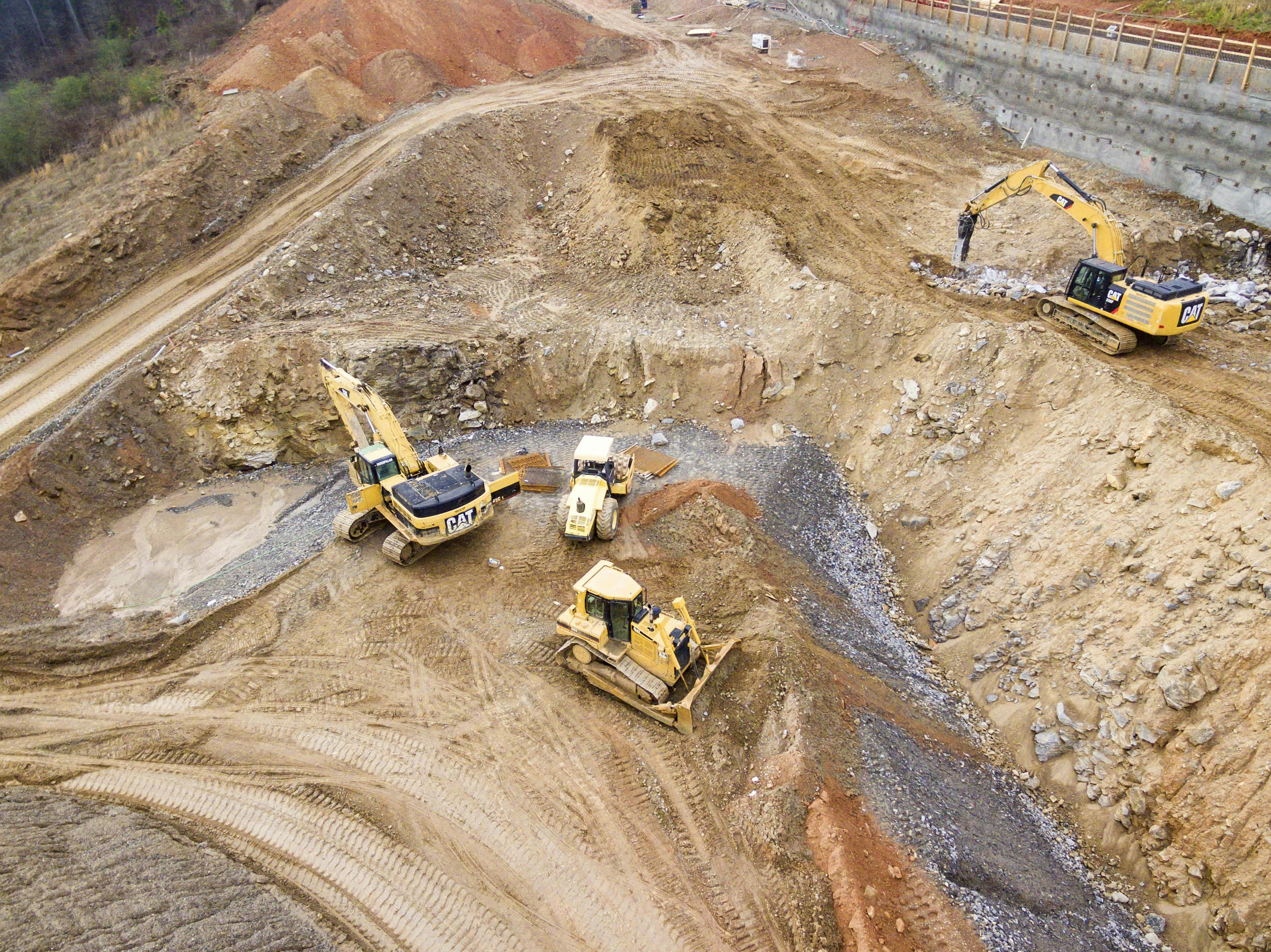 Construction worker in safety gear stands near large excavator moving rocks at construction site.