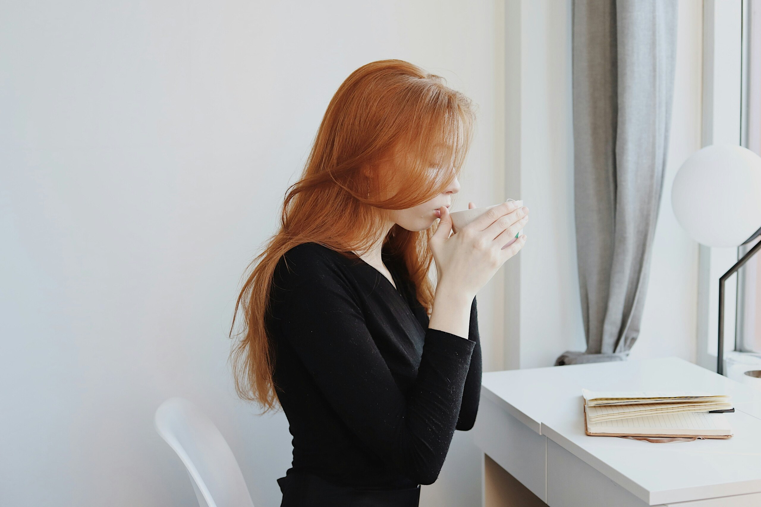 Mujer pelirroja bebiendo café junto a una ventana en un escritorio minimalista