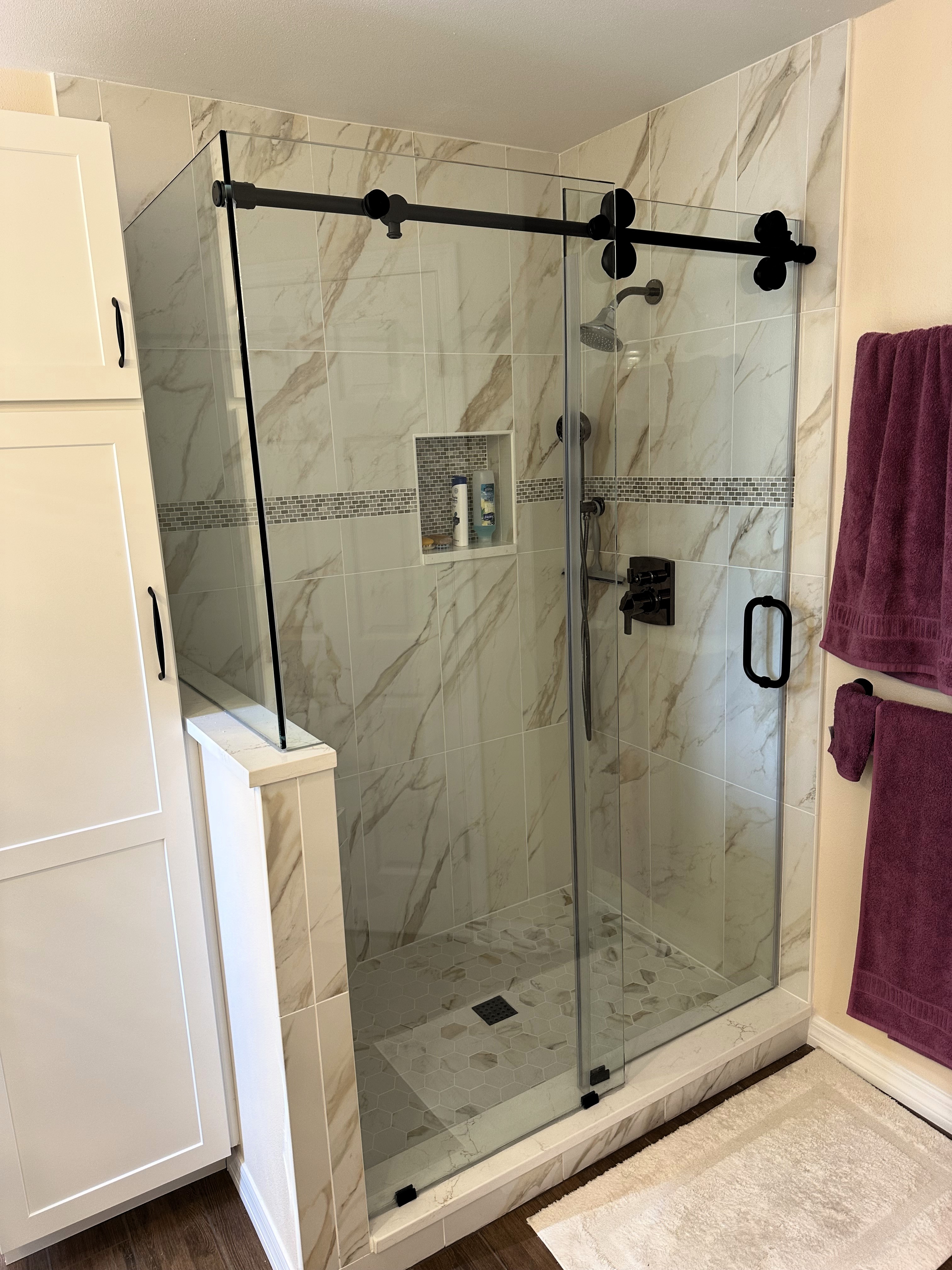 
This superb walk-in shower features a large barn-style rolling clear glass door with matte black hardware and
beautifully tiled 12x24 polished shower walls, hexagon mosaic honed shower floor, and decorative Mini-Brick band. 