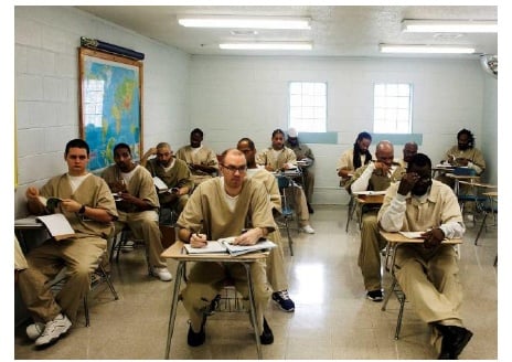 Inmates attending a class in a prison classroom setting, wearing beige uniforms and sitting at desks, with educational materials on the desks and a world map on the wall.