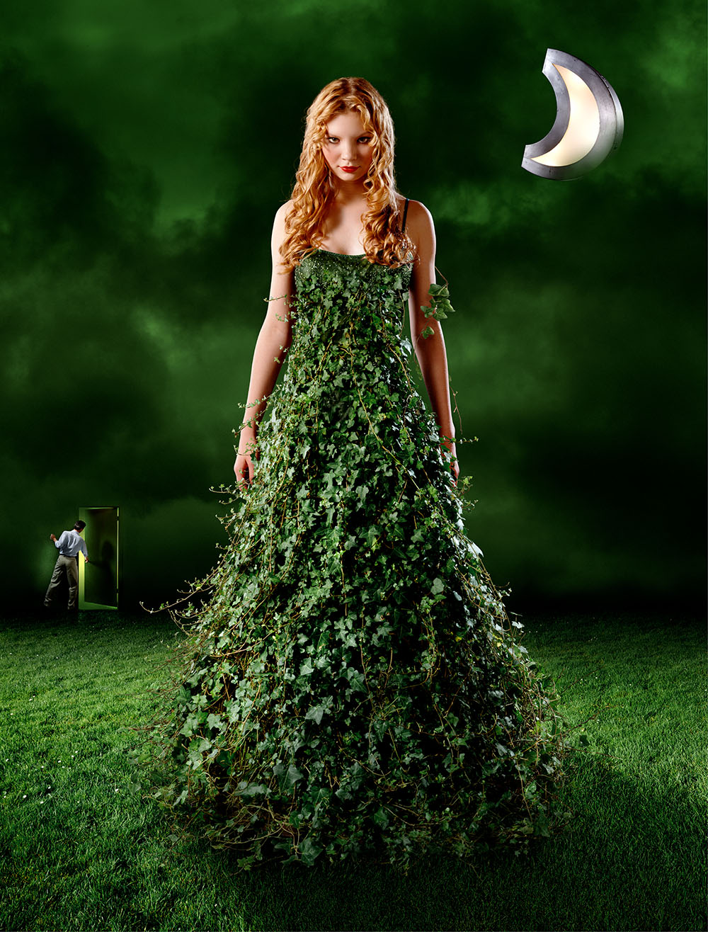 Surreal image of beautiful, red haired, woman wearing a dress made of ivy vines and leaves, standing in a field of green grass.