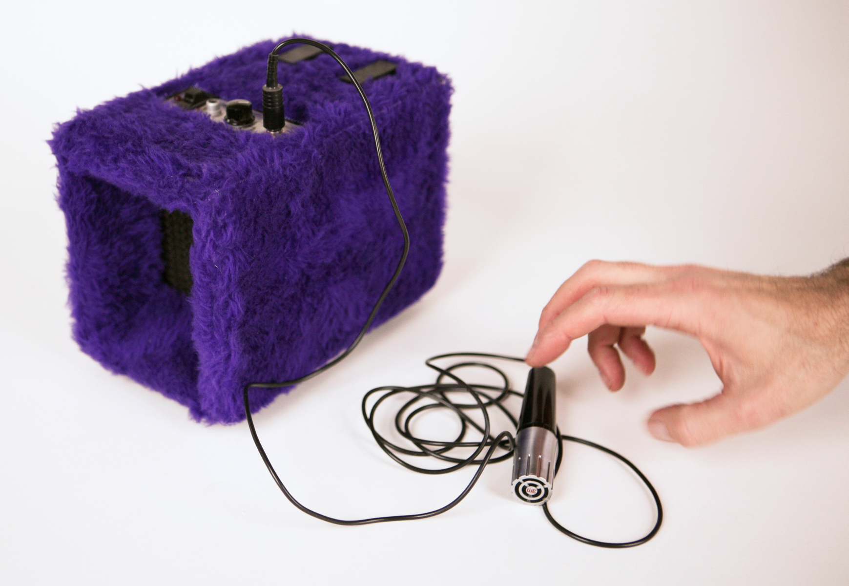 A hand reaches for a microphone plugged into a purple fur covered box device.