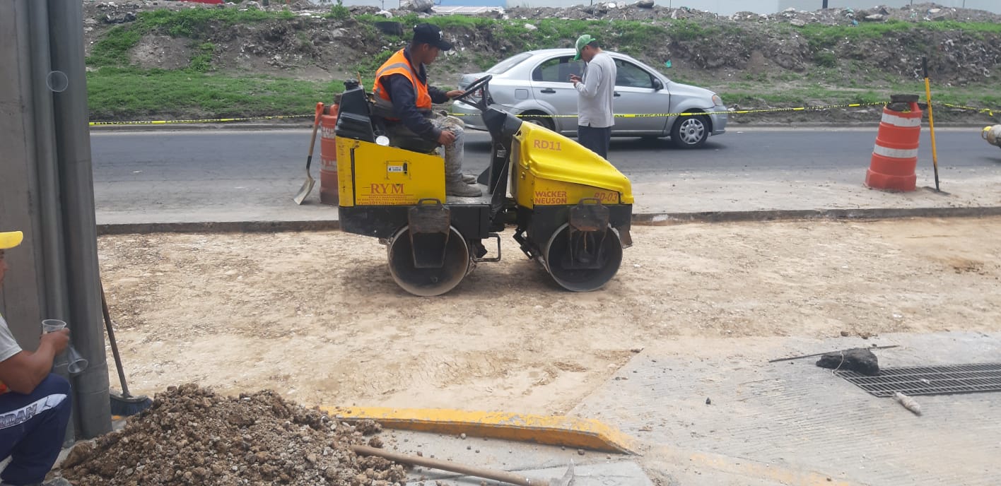 Obrero manejando una apisonadora pequeña en una construcción vial, con señalización de tráfico y otro trabajador al fondo. Obrero manejando una apisonadora pequeña en una construcción vial, con señalización de tráfico y otro trabajador al fondo.