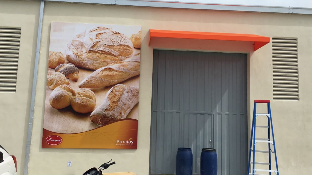 Fachada de edificio con cartel de panadería mostrando diferentes panes, escalera metálica azul y barriles azules junto a una puerta gris.