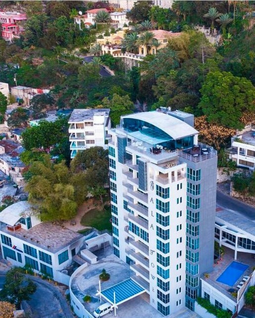 Vista aérea de un hotel moderno de gran altura con piscina, rodeado de árboles y viviendas en una zona urbana