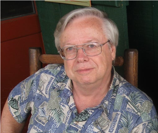 Elderly man sitting in a wooden chair, wearing glasses and a patterned shirt, with a pen in his pocket.